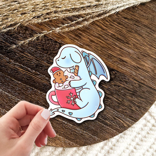 Cocoa Dragon waterproof vinyl die-cut sticker held in hand over a wood table, cute fantasy dragon decal gift for dragon fans and cozy winter lovers