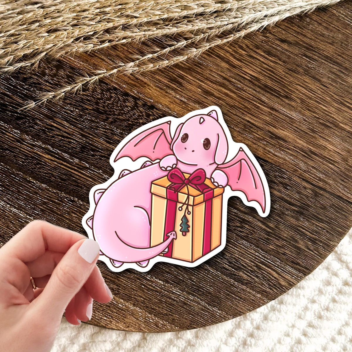 Gift Box Dragon vinyl die-cut sticker held in hand over a wooden table, cute pink baby dragon with red ribbon gift box, fantasy gift for dragon lovers