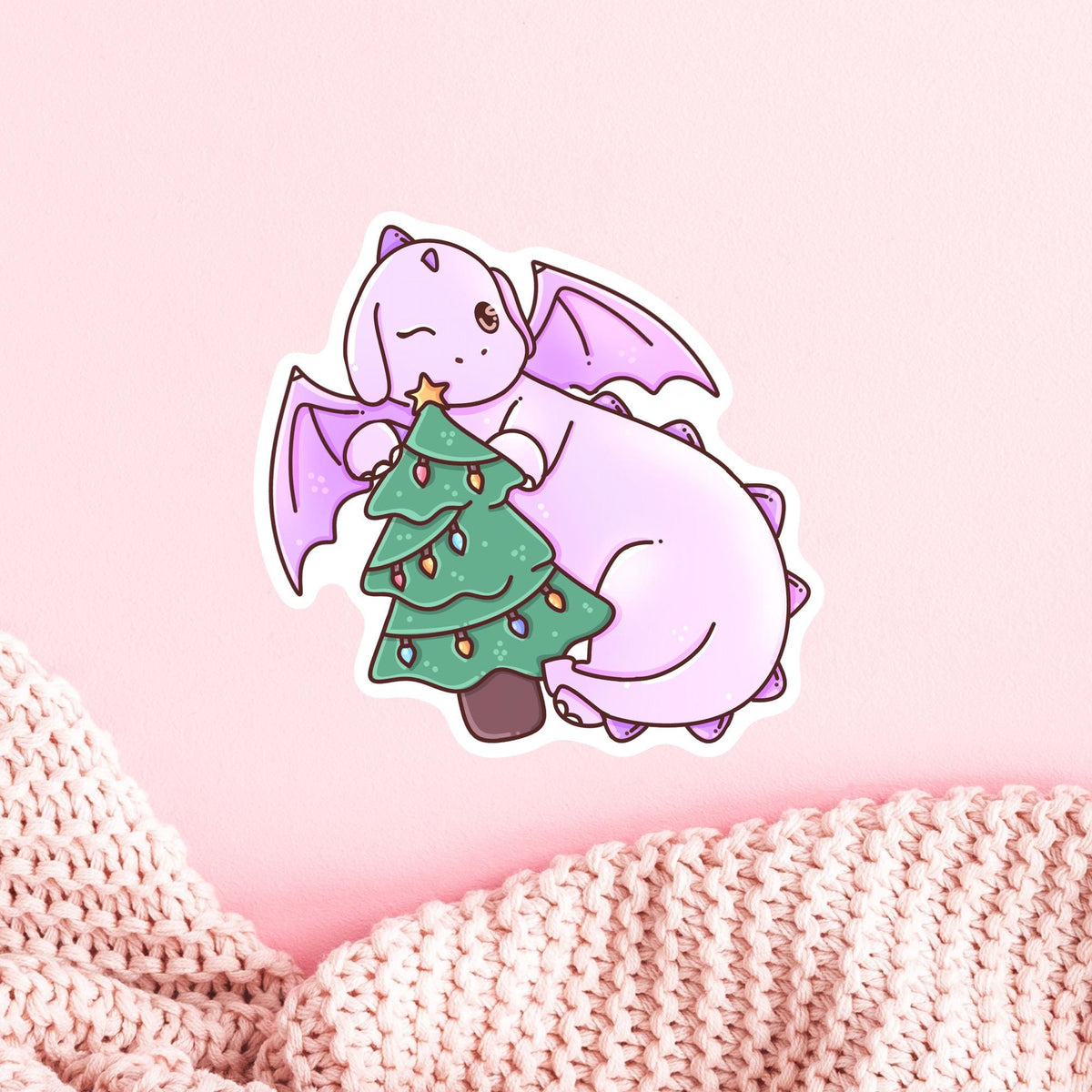 Christmas Tree Dragon die-cut vinyl sticker on a soft pink knit background, whimsical winter aesthetic stocking stuffer for dragon lovers