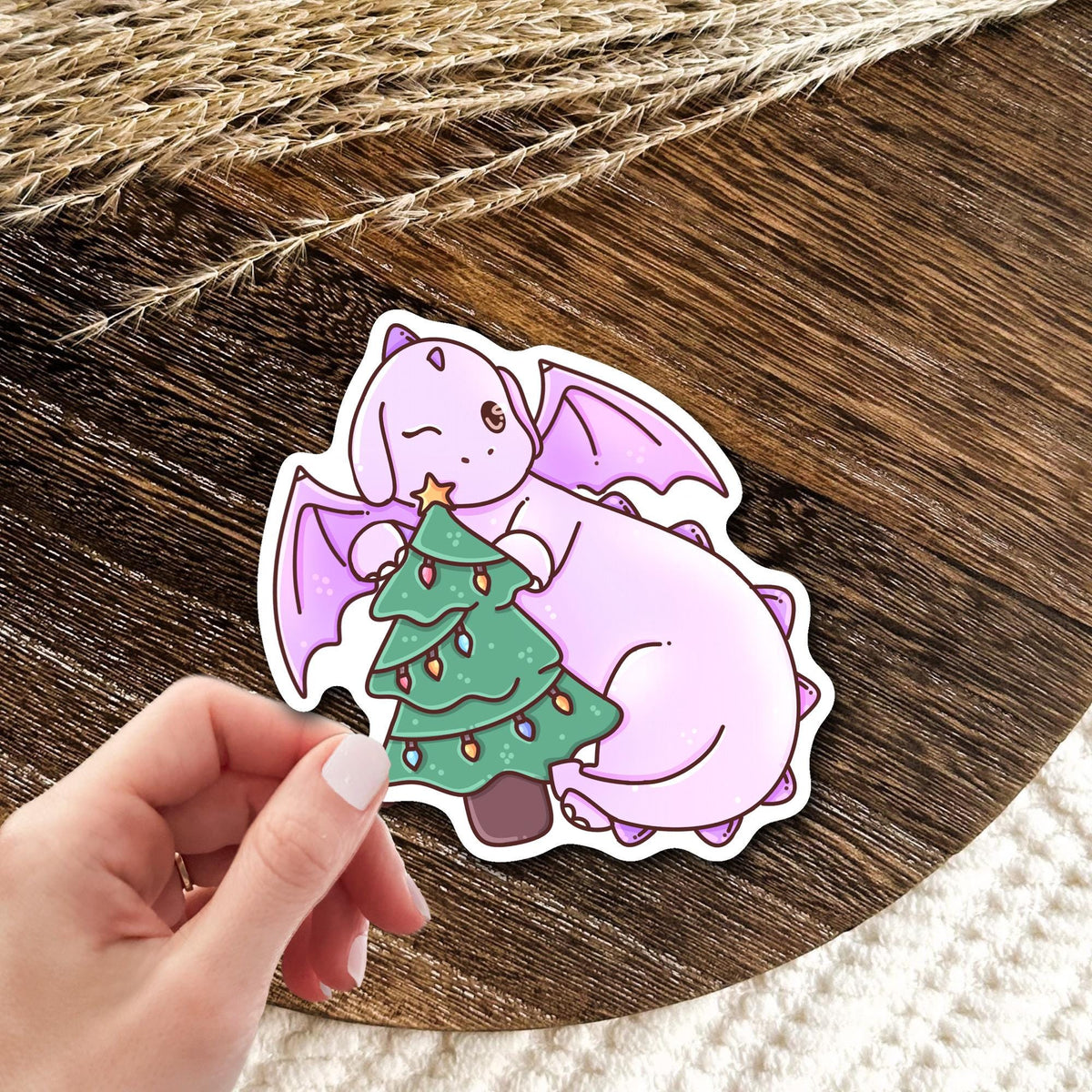 Christmas Tree Dragon vinyl die-cut sticker held in hand over a wooden table, cute holiday dragon sticker gift for fantasy lovers and Christmas fans