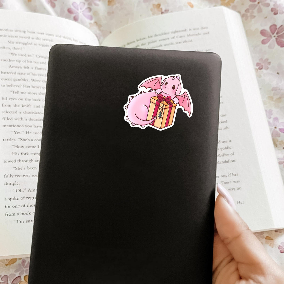 Gift Box Dragon vinyl sticker on a black phone case held over an open book, cute waterproof phone case decal for fantasy lovers