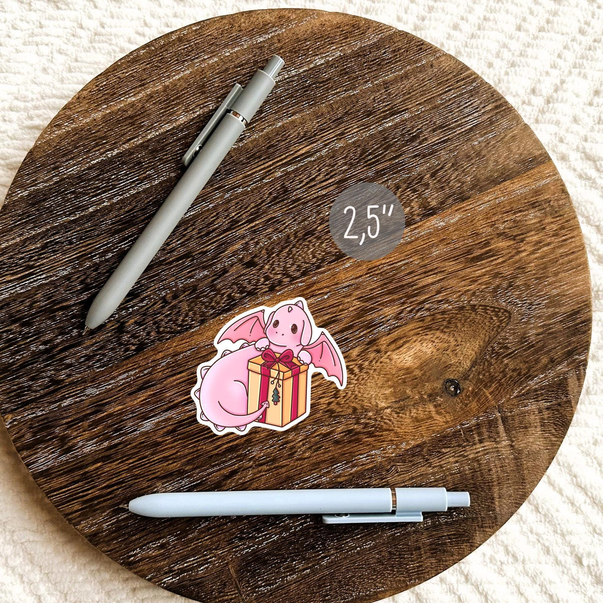 Gift Box Dragon sticker on a round wooden coaster next to a pen, cute fantasy vinyl decal for planners, journals and desks