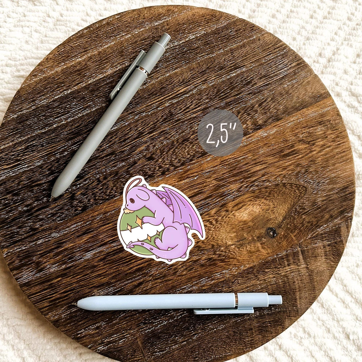 Small purple Ornament Dragon sticker on a round wooden tray next to a pen, waterproof vinyl journal sticker for notebooks and cozy holiday stationery