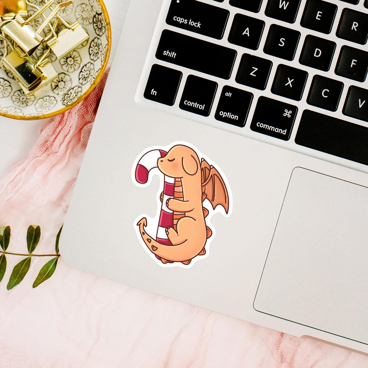 Candy Cane Dragon vinyl sticker on a round Candy Cane Dragon waterproof sticker on the back of a black e-reader or Kindle, cute holiday fantasy decal for e-readers, phone cases and tabletswooden desk with pens and notebook, waterproof Christmas sticker for journals, planners and cozy desk decor