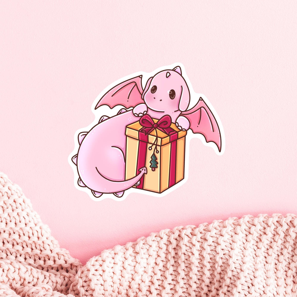 Pink baby dragon hugging a gift box, Gift Box Dragon die-cut vinyl sticker on a cozy pink knit background, whimsical winter gift idea