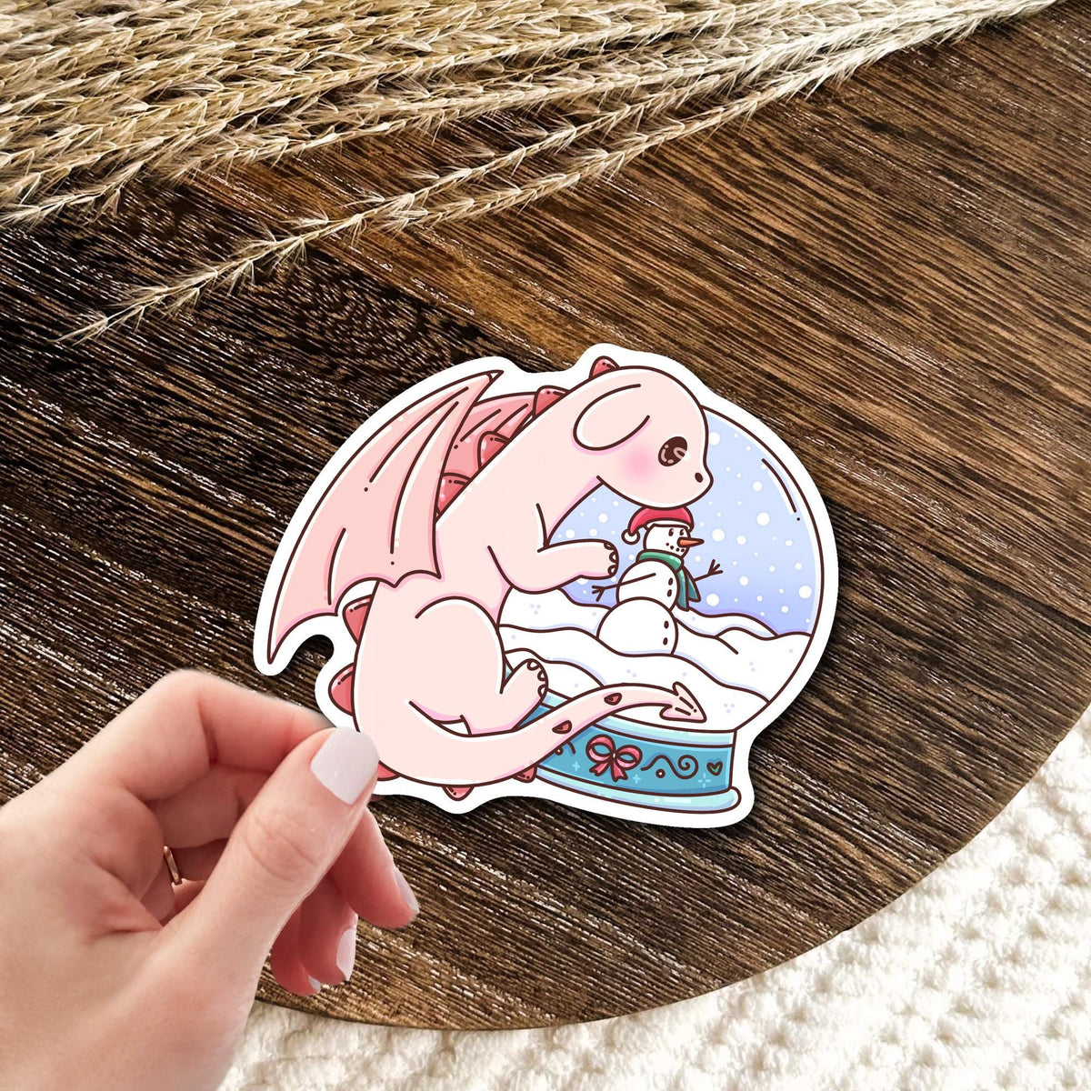 Snow Globe Dragon vinyl die-cut sticker held in hand over a wooden table, cute winter fantasy waterproof sticker gift for dragon lovers