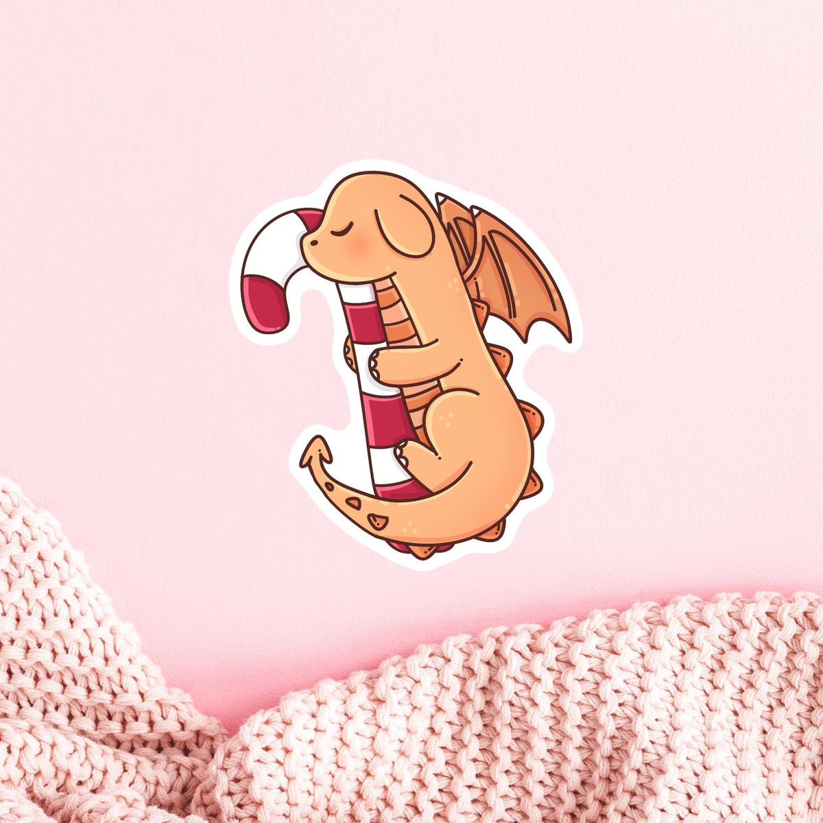 Cute Candy Cane Dragon die-cut sticker on a soft pink knit blanket, waterproof vinyl Christmas sticker in matte, glitter or holographic finish