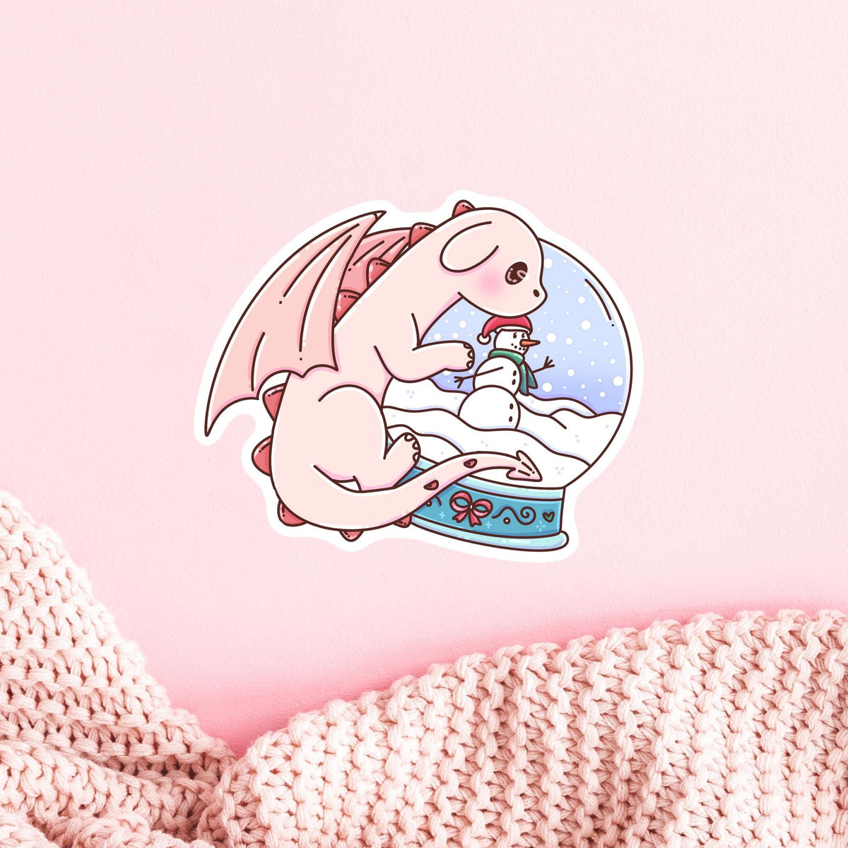 Cute Snow Globe Dragon die-cut vinyl sticker on a soft pink knit blanket, kawaii winter decal in matte, glitter or holographic finish